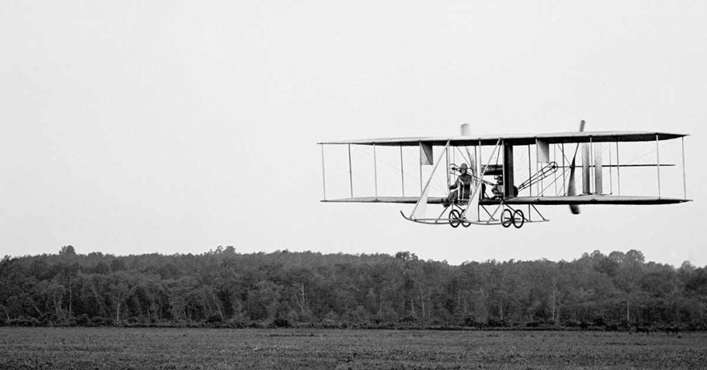 A monochrome photograph of an old-fashioned airplane soaring above a rural area.