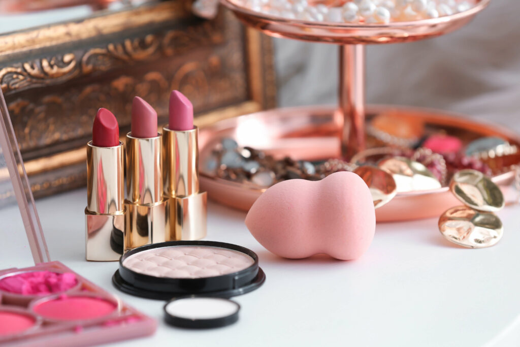 A makeup setup featuring three lipsticks in gold tubes, a powder compact, pink blush palette, and a pink makeup sponge sits elegantly beside a tray of assorted jewelry on a white surface, perfectly illuminated by the natural light streaming through the nearby glass door.