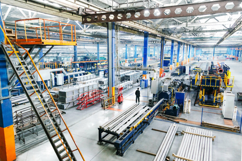 A spacious, well-lit factory floor with various industrial machines, metal stacks, and workstations. A worker stands in the center, overseeing operations through a glass door that separates different sections.