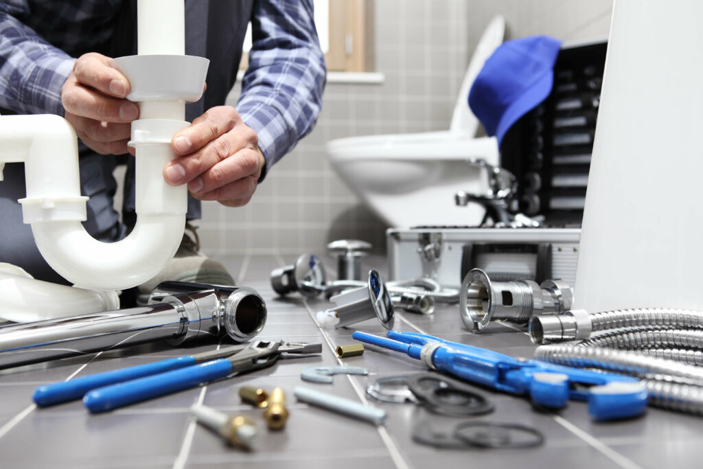 A person is fixing a pipe under a sink in a bathroom, with various plumbing tools and parts scattered on the tiled floor around them, much like the meticulous setup required in a plastic surgery business.