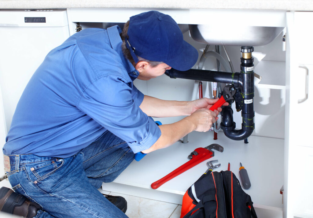 A plumber in a blue shirt and cap uses a wrench to fix pipes under a kitchen sink. Tools and a red tool bag are distributed on the floor beside him.