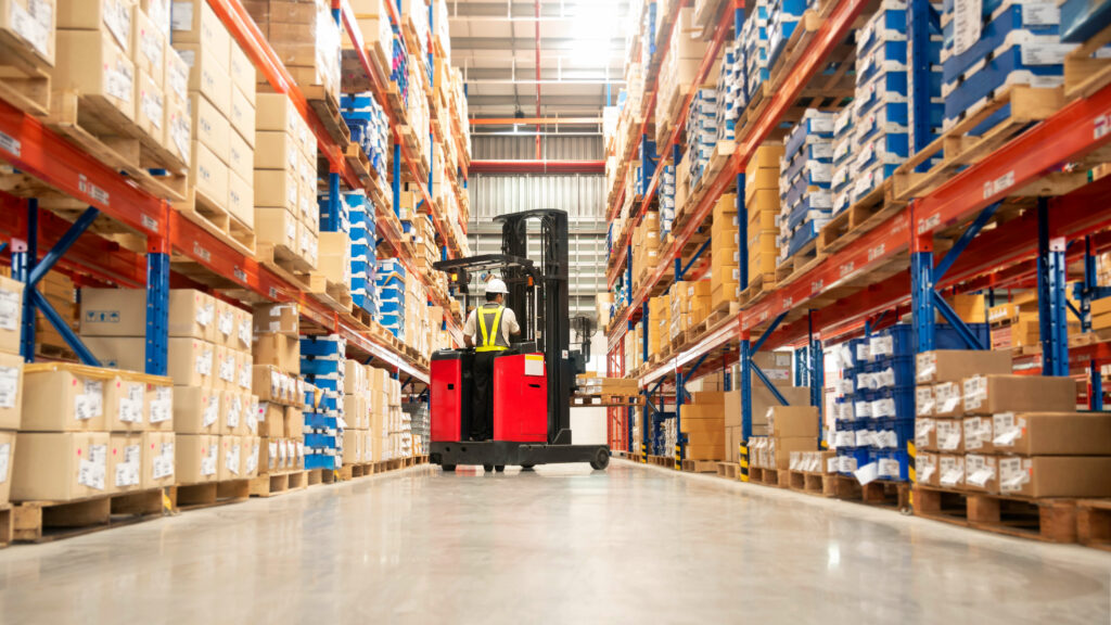 A worker operates a red forklift in a large warehouse aisle, surrounded by rows of stacked boxes and shelves, efficiently navigating the logistics of the supply chain.