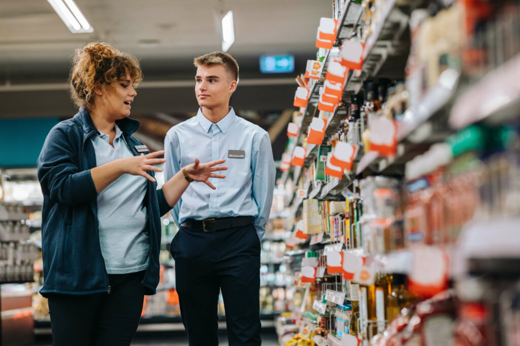 Two people in professional attire stand in a grocery store aisle, discussing products on the shelves. One gestures while explaining the importance of garment care products, and the other listens attentively, as though they were choosing between laundry services or dry cleaners for their business.