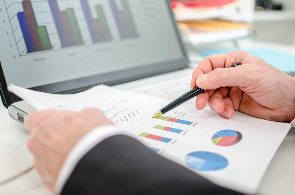 A person in business attire reviews printed charts and graphs with a pen in hand, analyzing Adjusted EBITDA figures, while similar charts are displayed on a laptop screen in the background.