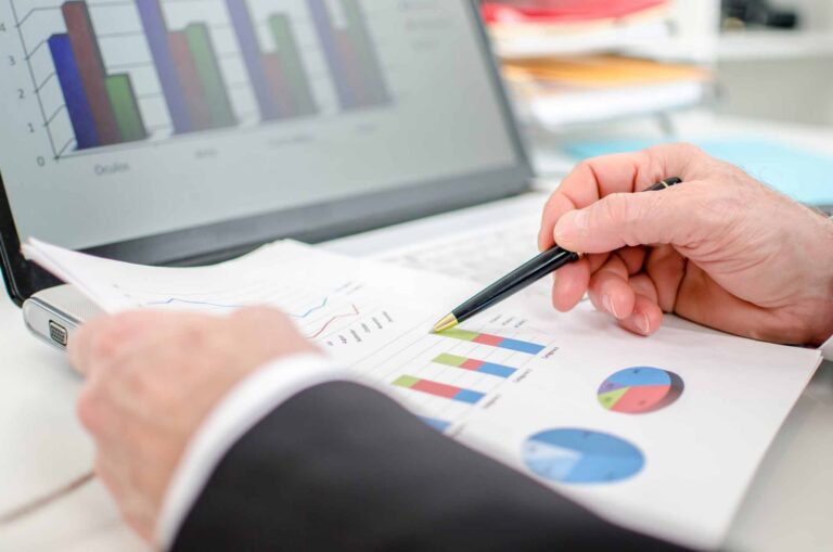 A person in business attire reviews printed charts and graphs with a pen in hand, analyzing Adjusted EBITDA figures, while similar charts are displayed on a laptop screen in the background.