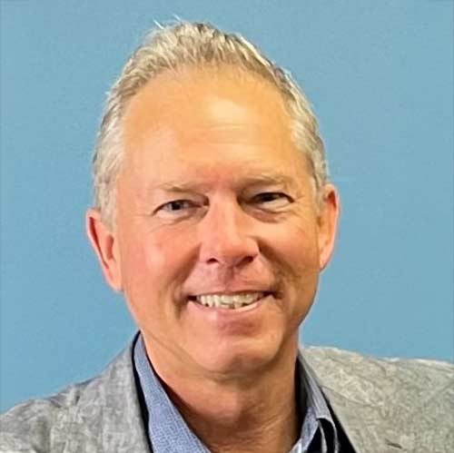 Craig Sweeney, with short gray hair, smiles at the camera against a blue background. He wears a gray jacket and blue shirt, exuding warmth and charm.