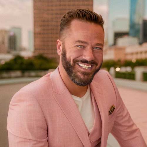 Jason Lavrack, a smiling man with a beard, confidently stands outdoors in his pink blazer and white shirt, the blurred city buildings providing an urban backdrop.