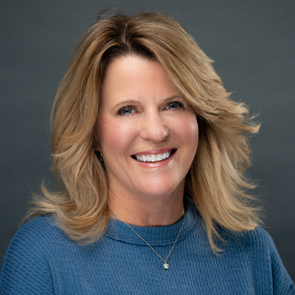 Smiling woman with shoulder-length blonde hair wears a blue sweater against a dark gray background.