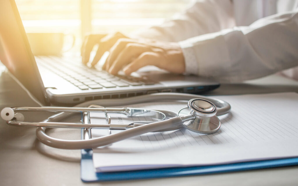 A doctor types on a laptop with a stethoscope and clipboard on the desk in soft lighting.
