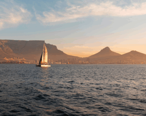 A sailboat glides across calm water with a city and mountains, including Table Mountain, in the background at sunset—an inspiring scene for business owners seeking essential insights on selling your business.