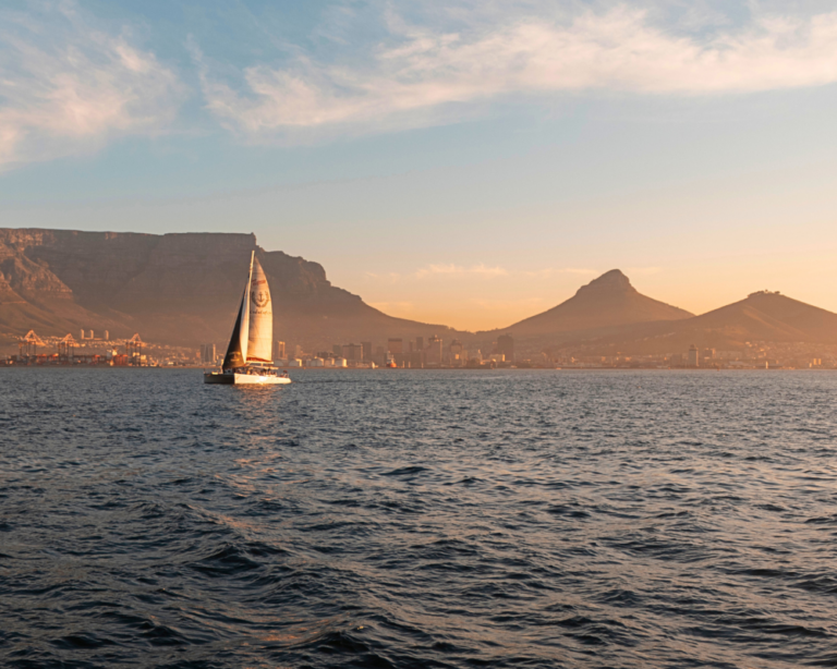 A sailboat glides across calm water with a city and mountains, including Table Mountain, in the background at sunset—an inspiring scene for business owners seeking essential insights on selling your business.
