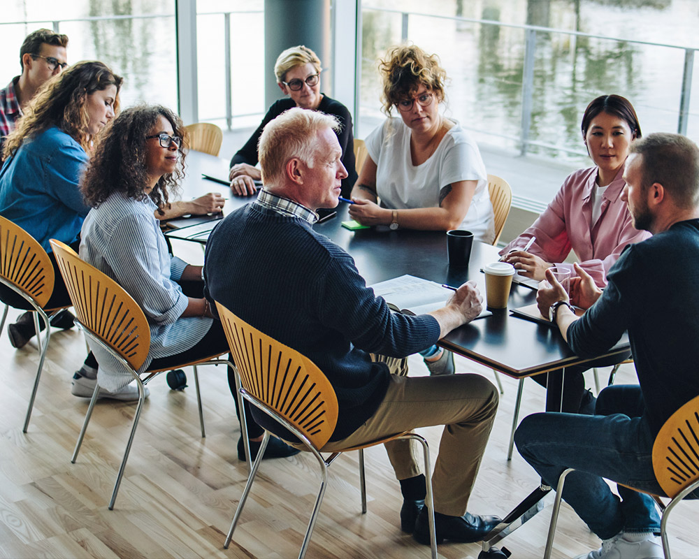 A group of eight people sit around a table in a modern office, engaged in a discussion about how to sell your business, with papers, notebooks, and coffee cups in front of them.