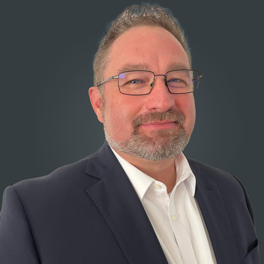 A man with glasses, short hair, and a trimmed beard wearing a dark suit jacket and white shirt, standing in front of a plain dark background.