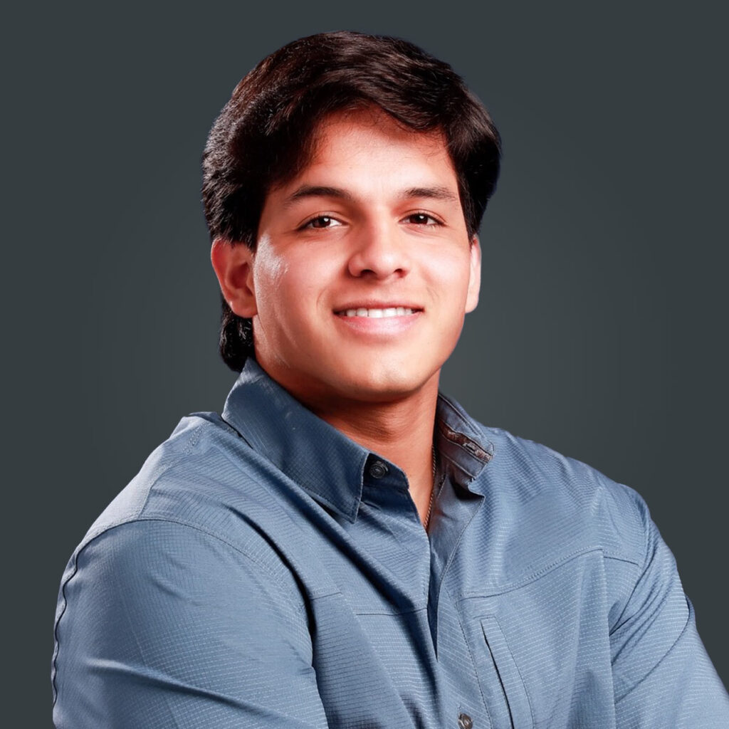 A young man with dark hair wearing a blue button-up shirt smiles at the camera against a plain dark background.