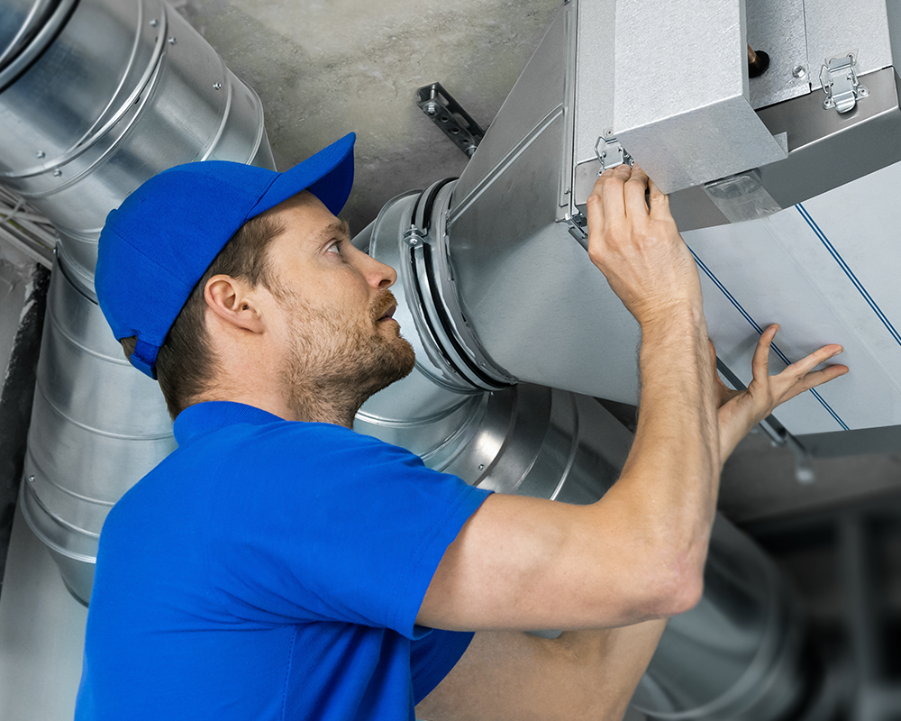 A man in a blue uniform and cap adjusts or inspects a metal air duct or ventilation system on a ceiling.