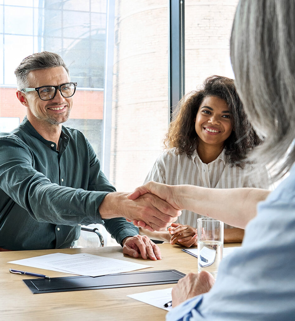 Three people meet at a table; two, a man and a woman, smile as the man shakes hands with a third person whose back is to the camera. Documents and a glass of water suggest a merger and acquisitions advisory meeting.